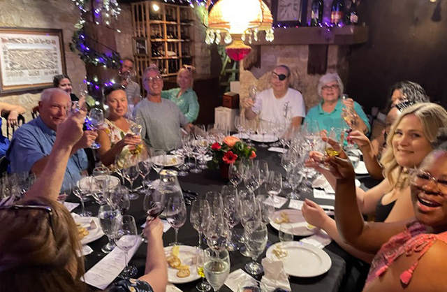 Happy group seated at wine tasting table raising glasses to the camera.